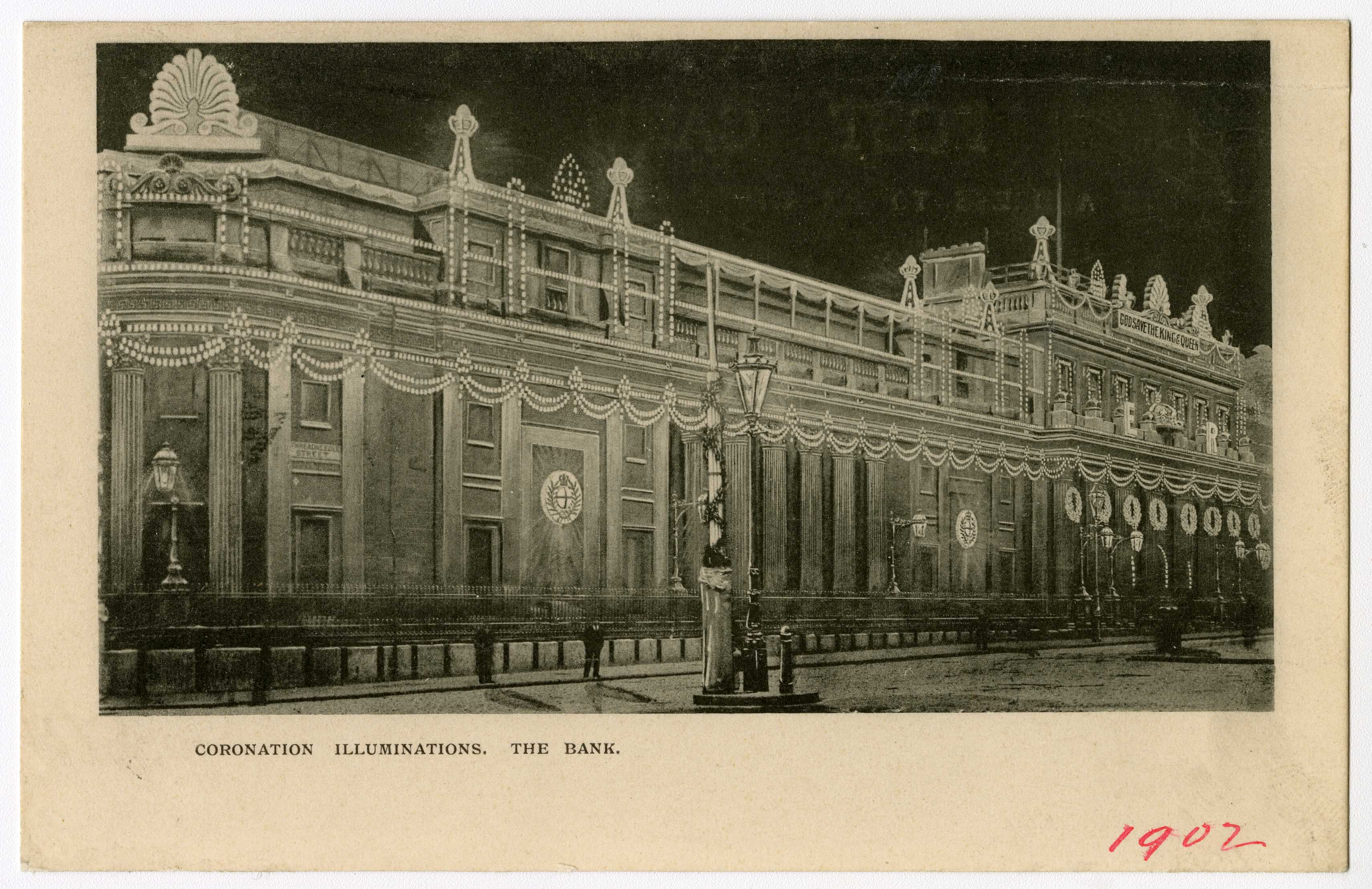 A black and white photograph of the Bank of England at Threadneedle Street in 1902, at night. The bank is decorated with banners and the initials E R, for the coronation of Edward VII. Most of the scene is in darkness but the illuminated banners and strings out lights show up clearly. 
