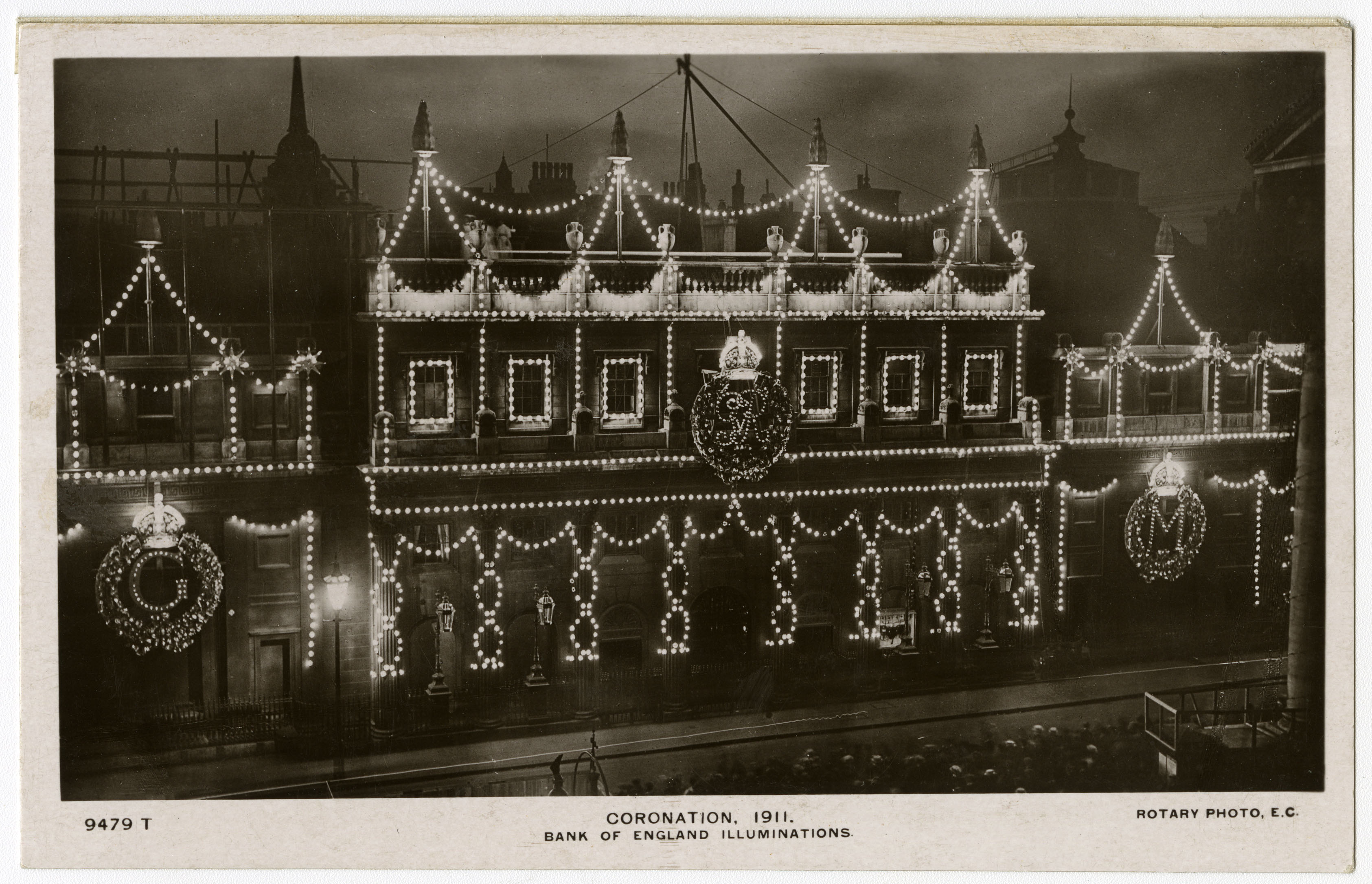 A black and white photograph of the Bank of England at Threadneedle Street at night. The Bank is decorated with strings of lights for the coronation of George V in 1911. The illuminated banners and strings out lights show up clearly against the darkness.