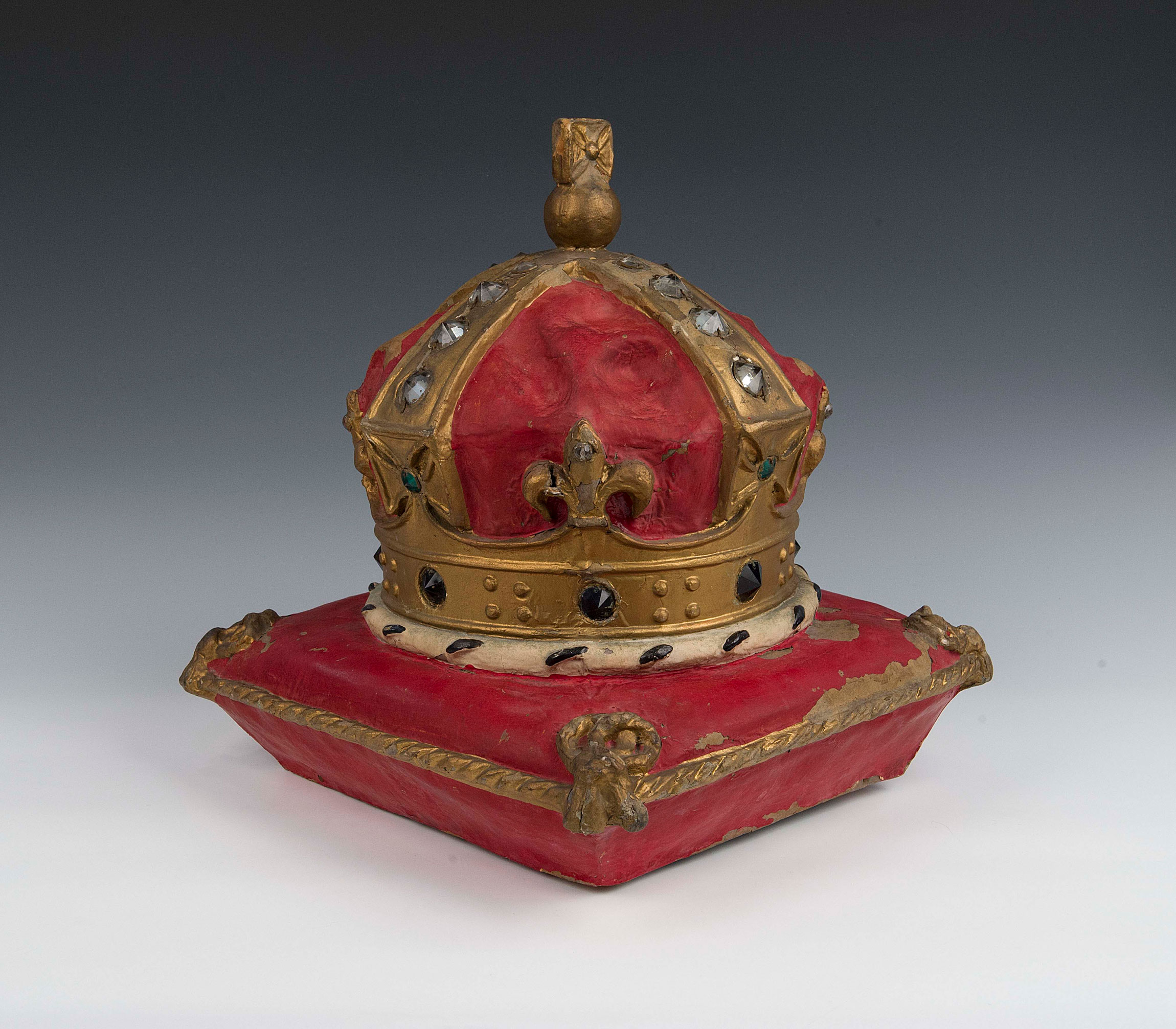 A painted model of a gold crown sitting on a red cushion that has golden tassels. The jewels on the crown are made of glass, which shine when the model is lit up from the inside. 