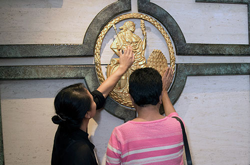 Audio described tour CTA a member of Bank staff and a partially sighted visitor stand in front of a large, round, gold, metal relief of the female figure of Brittania, symbol of the Bank of England. Both are tracing the shape with their hands